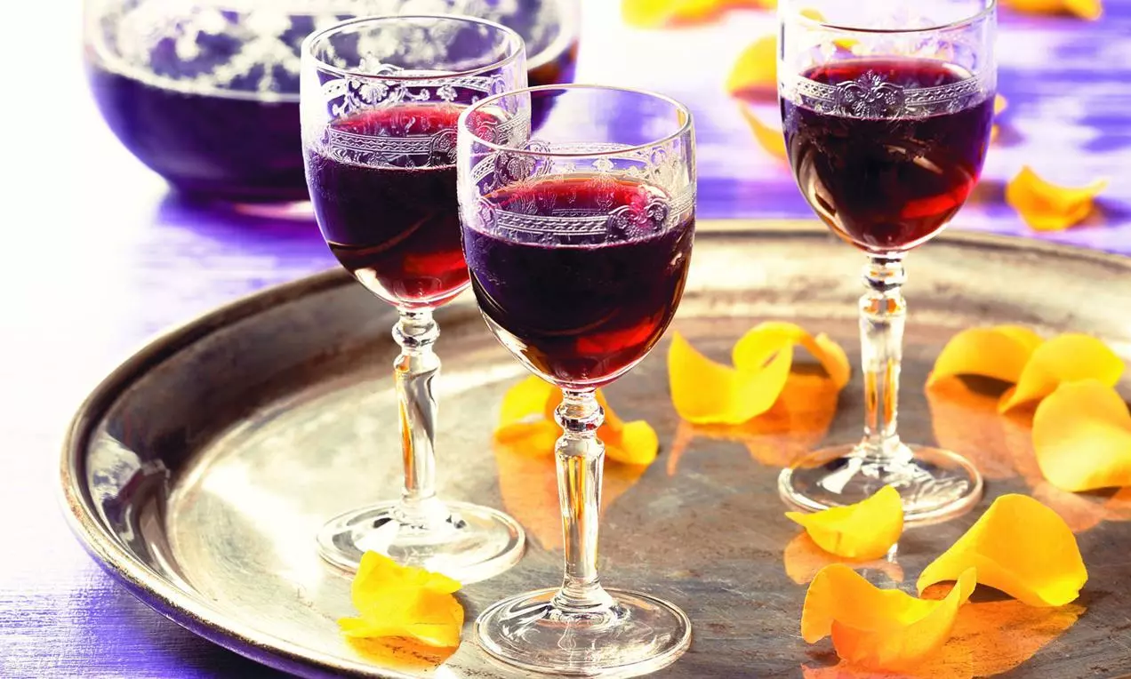 Tre calici di liquore rosso rubino su un vassoio argenteo, decorati con petali giallo vivo sparsi su fondo viola luminoso