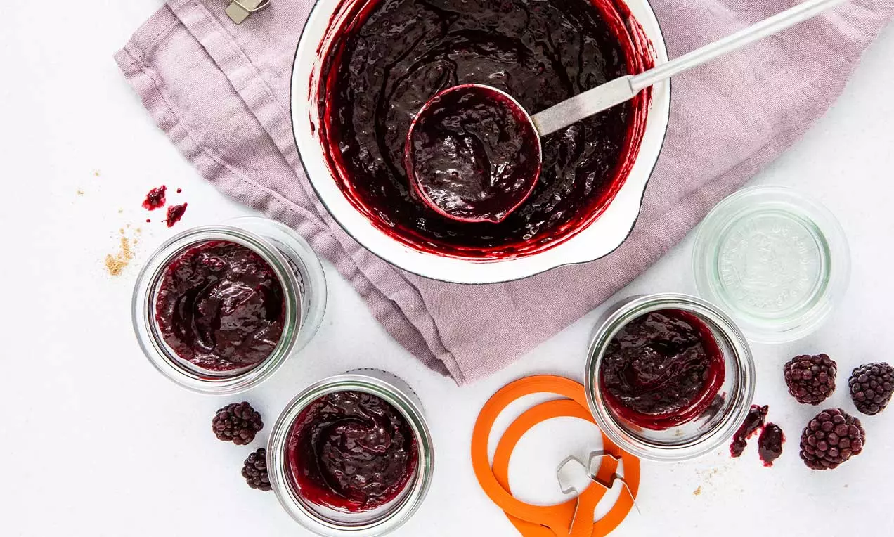 Dunkelviolette Brombeer-Konfitüre mit glatter, glänzender Oberfläche in offenem Topf und Gläsern auf einem fliederfarbenen Tuch arrangiert