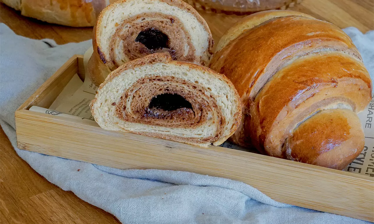 Halbiertes Zebra-Croissant mit dunkelbrauner Kakaoschicht inmitten goldbraunen Teigs in einem Holztablett auf hellem Stofftuch drapiert