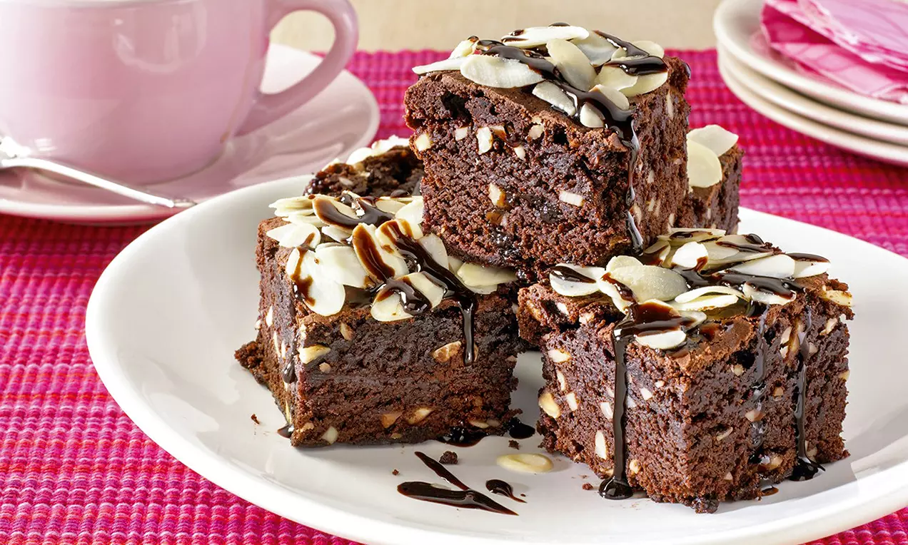 Cubes de gâteau au chocolat brun foncé parsemés d’amandes effilées et nappés de filet de chocolat sur une assiette blanche
