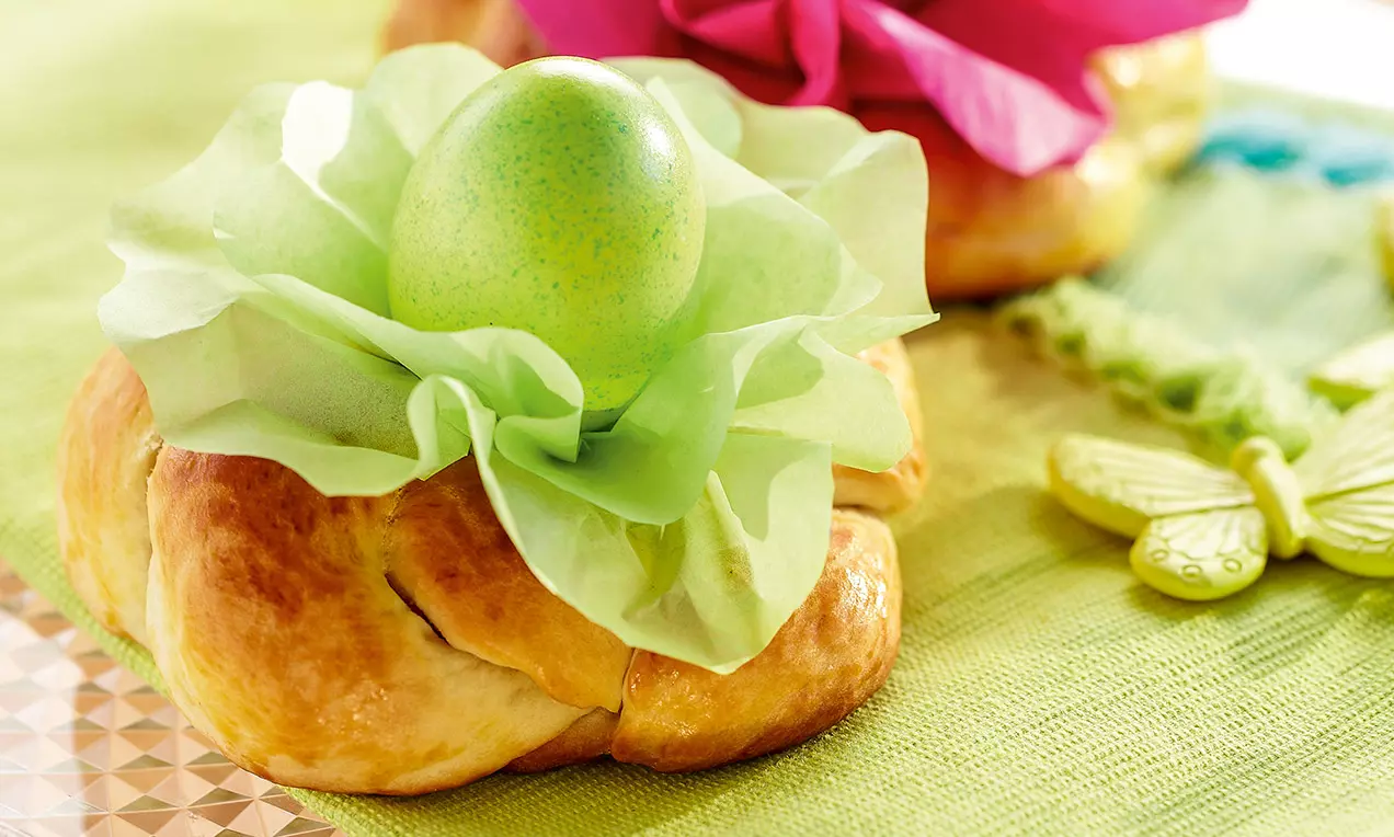 Panetti dorati intrecciati con crosta lucida e uovo verde posato su veli di carta verde, riposti su tovaglia verde con decorazioni
