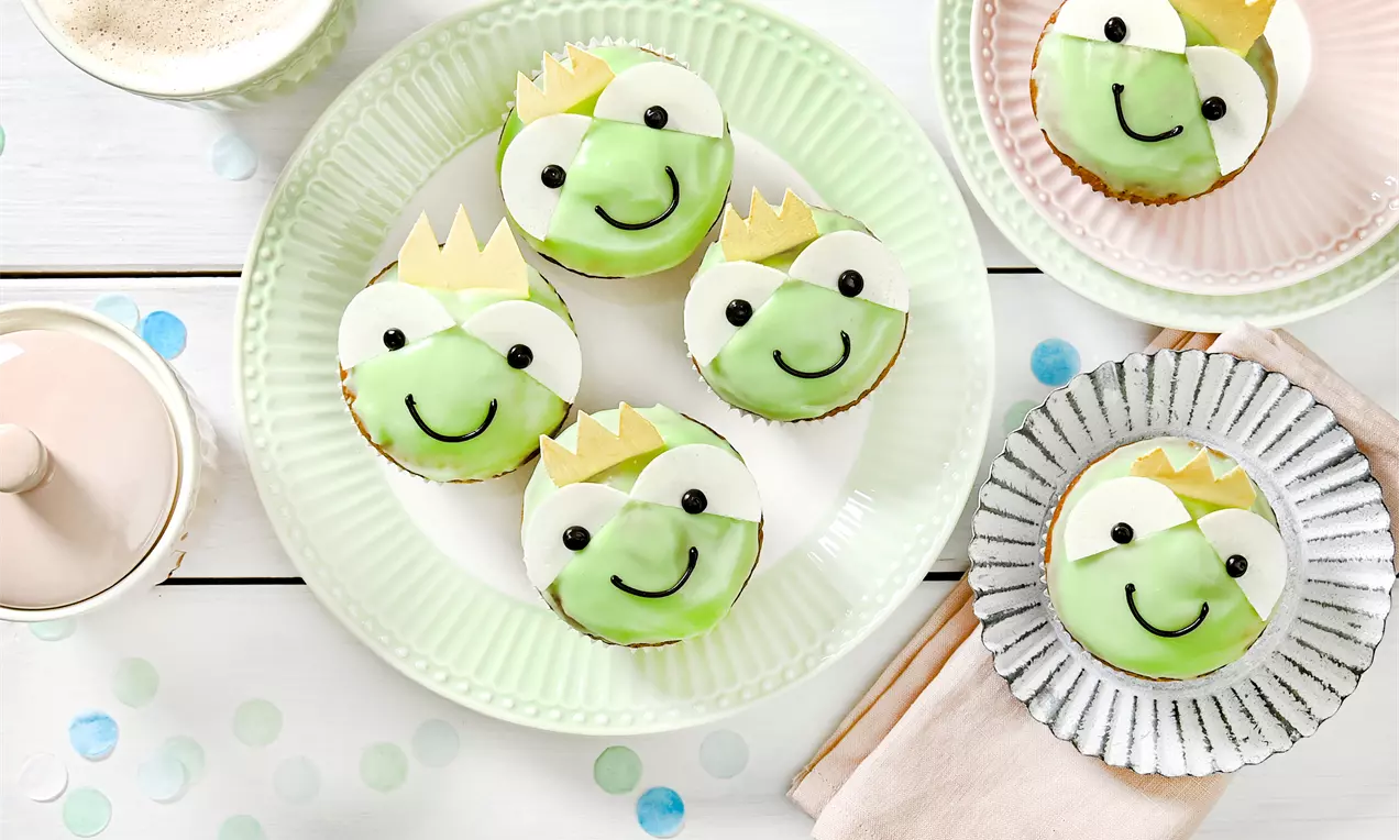 Muffin decorati con facce di rana verde chiaro, occhi bianchi e sorrisi neri, coronati da piccole stelle dorate su piatti pastello