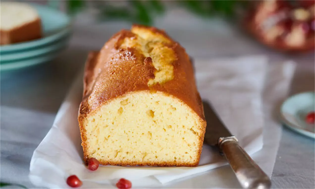 Heller, rechteckiger Rührkuchen mit goldbrauner Kruste und lockerer Textur auf weißem Pergamentpapier, umgeben von Granatapfelkernen
