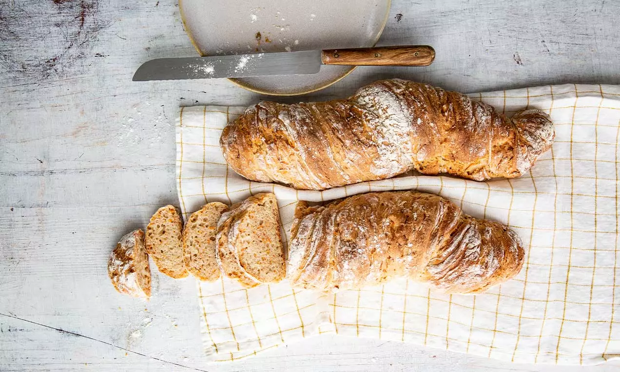 Pane di farina d'Urdinkel dorato e rugoso con carote, dalla forma rustica e leggermente attorcigliata, su un canovaccio a quadretti