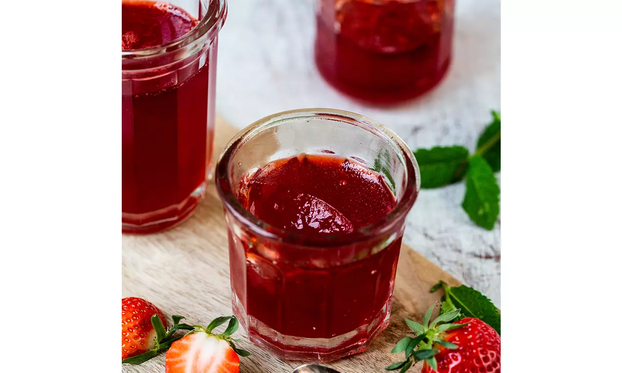 Gelée rouge translucide aux fraises dans un verre cannelé, entourée de fraises fraîches et de feuilles de mélisse sur une planche en bois