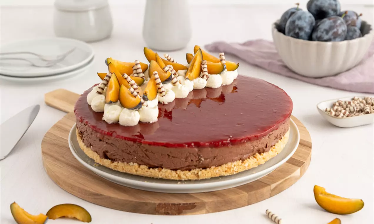 Schokoladen-Cheesecake mit glänzend rotem Zwetschgenbelag, Sahnetupfen, frischen Zwetschgen und Schokoröllchen auf hellem Keksboden.
