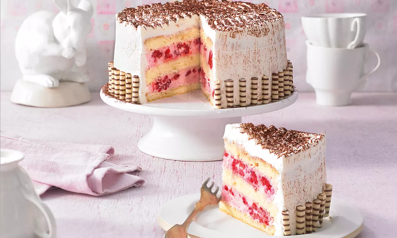 Kleine Himbeertorte auf weißer Platte mit Schokoröllchen umrandet, luftigem Biskuit, rosa Himbeerfüllung und Sahne, dekoriert mit Schokoraspeln