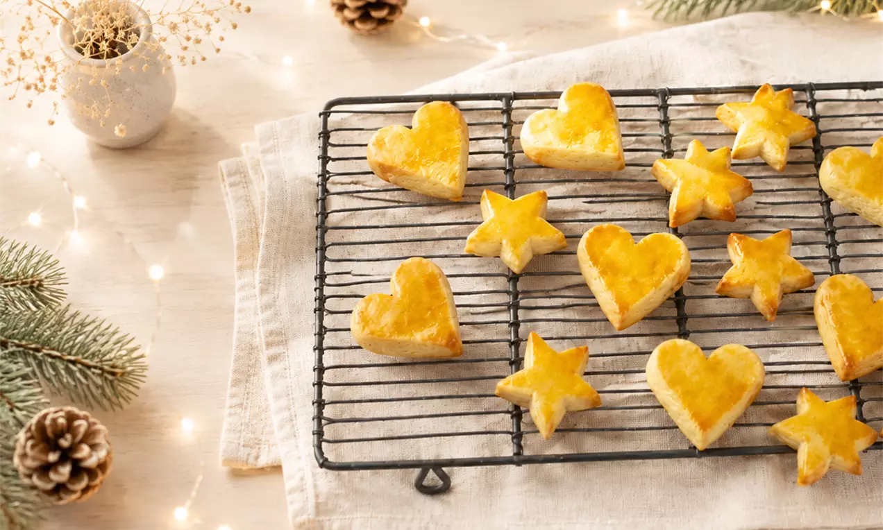 Biscuits Milanais dorés en forme de cœurs et d’étoiles disposés sur une grille noire, entourés de décorations hivernales., AI-labeled image
