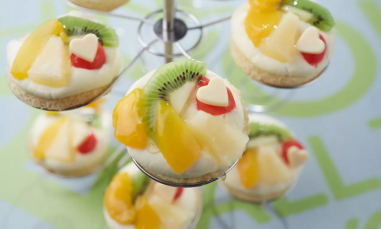 Petits fonds de tarte dorés garnis de crème vanille blanche, fruits brillants colorés et cœurs en chocolat, disposés sur un présentoir argenté