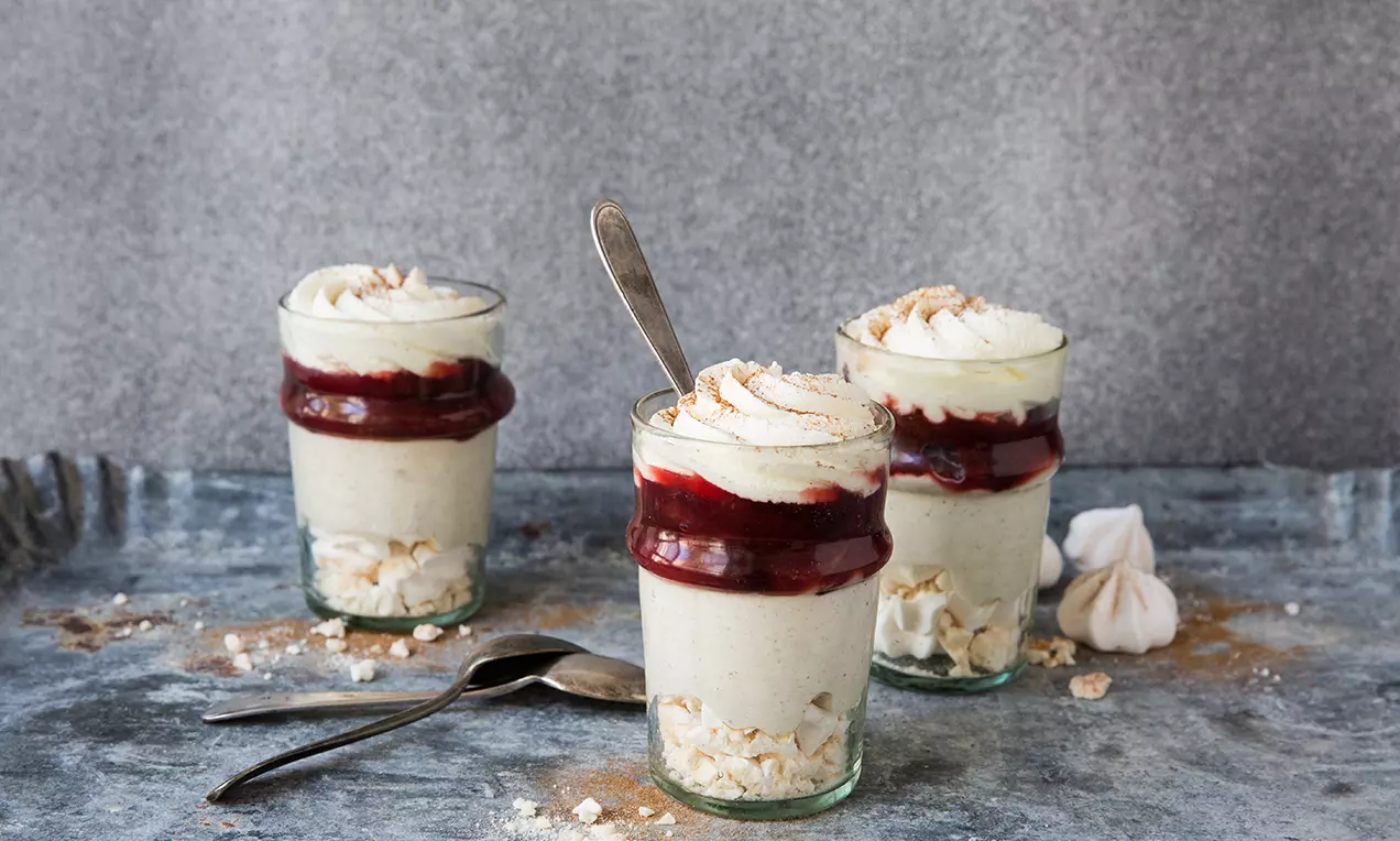 Mousse beige à la cannelle en verrines avec éclats de meringue, couche de coulis rouge et chantilly saupoudrée sur une table grise