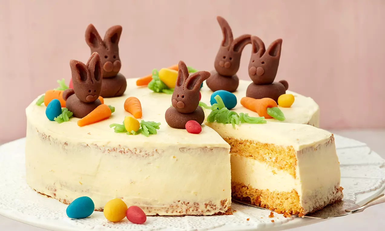 Orangen-Möhren-Torte mit heller Creme, dekoriert mit Schoko-Osterhasen, bunten Eiern und Modelliermöhren auf weißer Tortenplatte