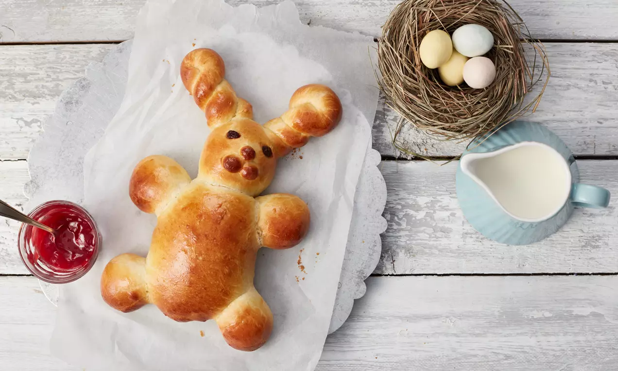 Goldbraunes süßes Hefeteighäschen mit glänzender Oberfläche auf Backpapier, daneben ein Nest mit bunten Eiern und hellblauer Milchkanne