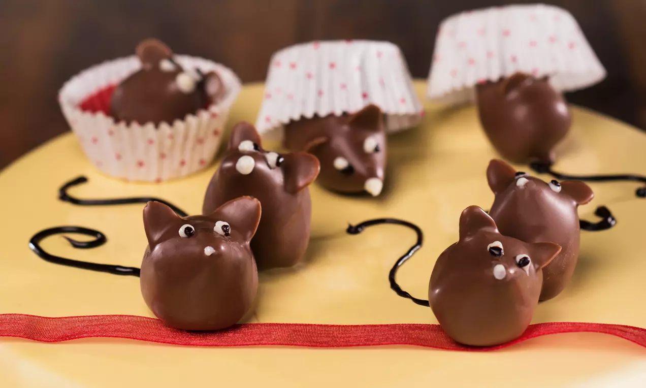 Dolci truffle a forma di topolini marroni, con occhi bianchi e code di cioccolato su un piatto giallo decorato con nastri rossi e pirottini bianchi
