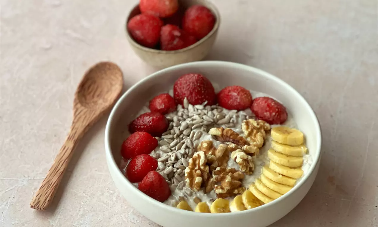 Cremiges Müesli in einer weißen Schale, belegt mit roten Erdbeeren, gelben Bananenscheiben, Walnüssen und hellen Sonnenblumenkernen