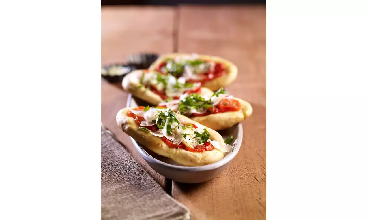 Goldgelbe oval geformte Pizza-Häppchen mit rotem Tomatenbelag, geschmolzenem Käse und frischen grünen Kräutern auf weißer Keramikschale