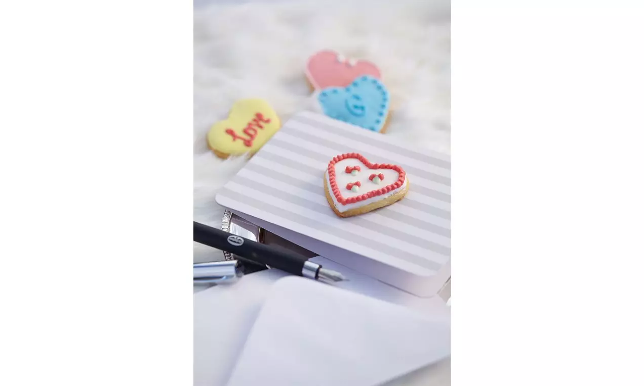 Biscotti a forma di cuore decorati con glassa colorata e dettagli raffinati, disposti su una scatola a righe su fondo chiaro e morbido
