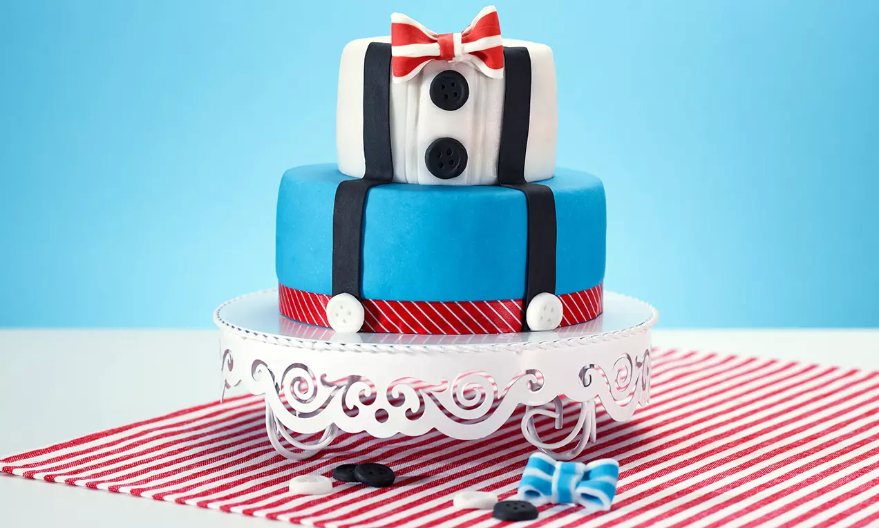 Torta a due piani con copertura azzurra e bianca, decorata con bretelle nere, bottoni e papillon rosso su alzata bianca traforata
