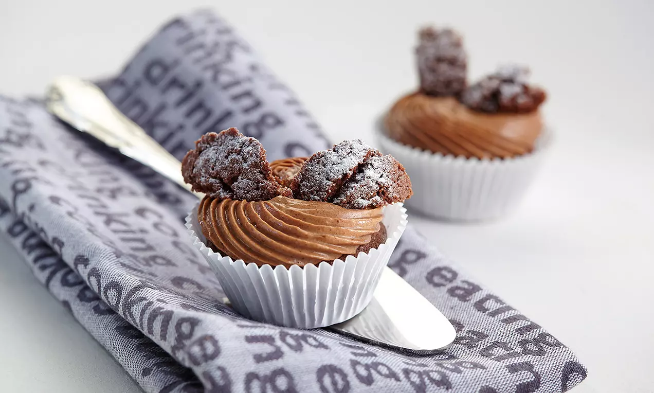 Petits cupcakes chocolatés dans des caissettes blanches, garnis de crème ridée couleur cacao et décorés de morceaux saupoudrés de sucre neige