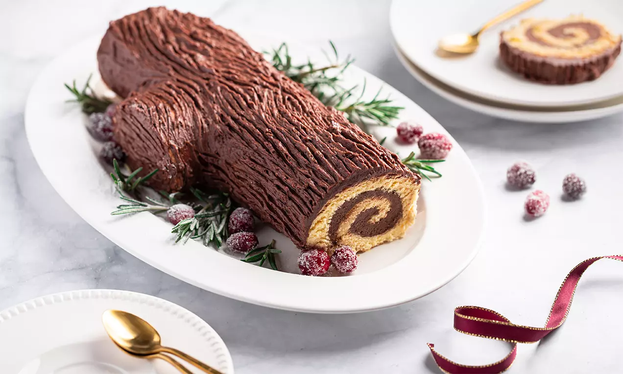 Bûche de Noël au glaçage brun texturé façon écorce, rouleau doré visible à l’intérieur, décorée de romarin et baies rouges sur un plat blanc