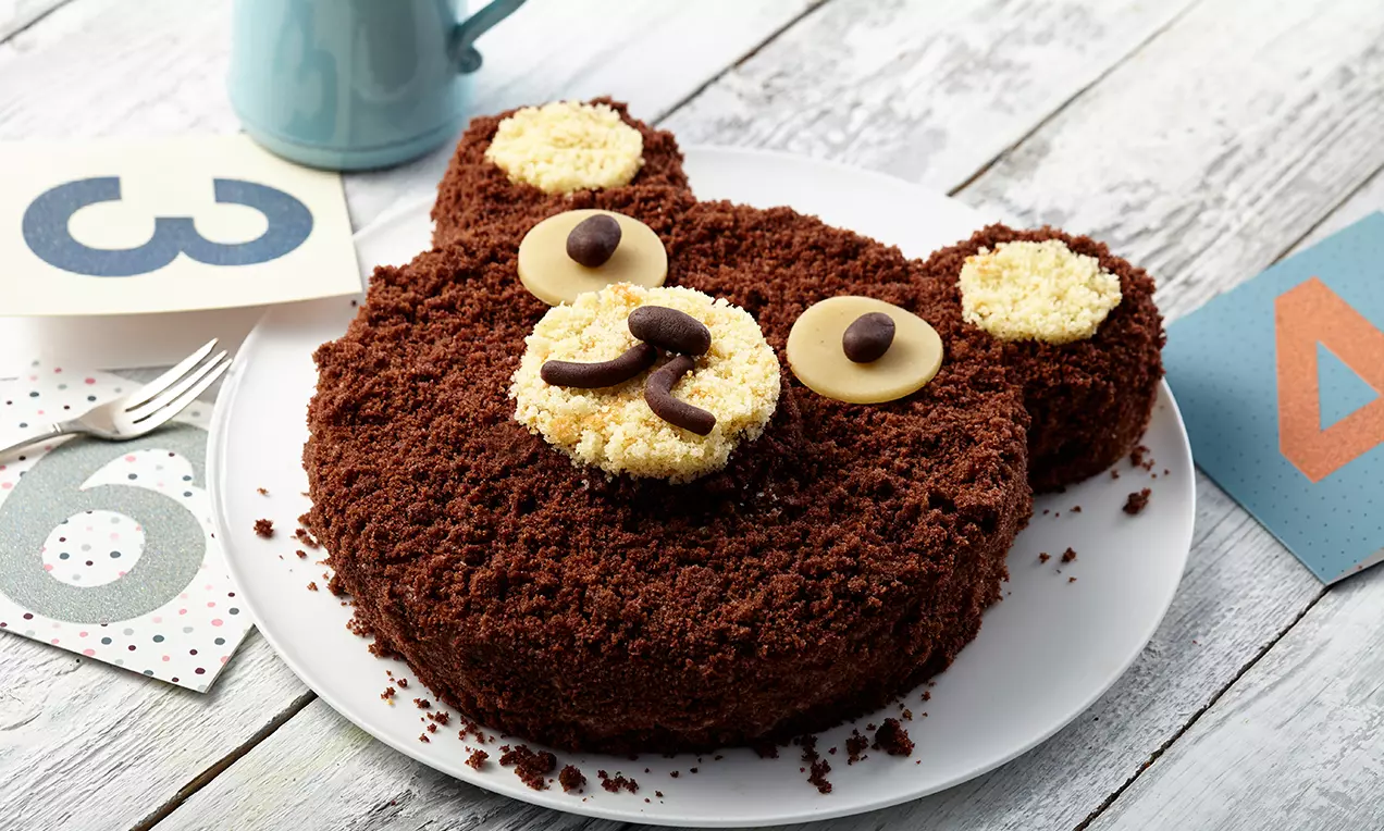 Gâteau en forme de tête d'ours avec une texture chocolatée foncée, oreilles et museau garnis de biscuit, yeux en chocolat sur une assiette blanche