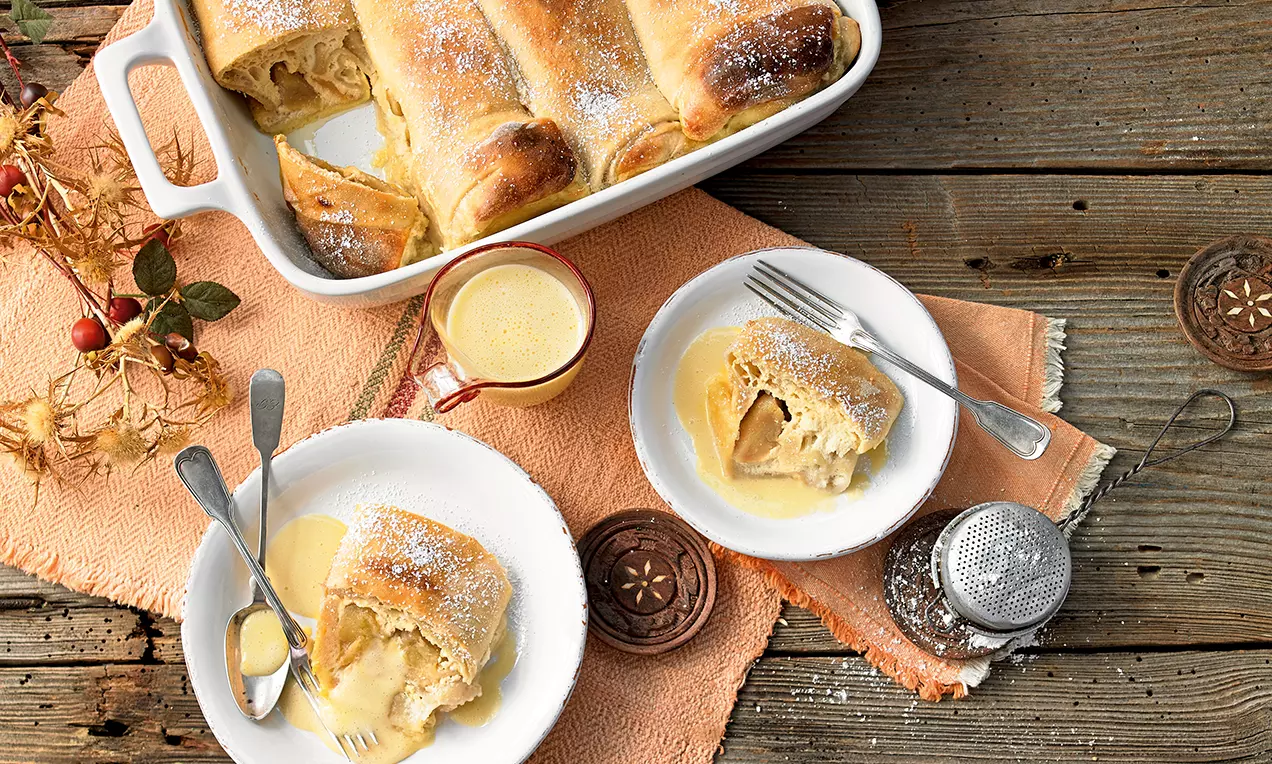 Milchrahmstrudel goldbraun mit feiner Füllung, karamellige Äpfel und Vanillesauce auf Teller angerichtet, in Schale und auf Holztisch serviert