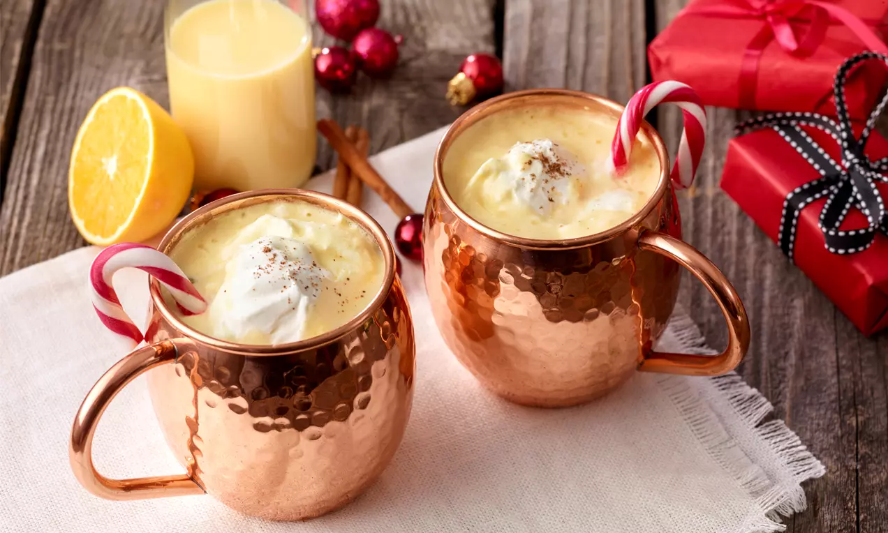Punch crémeux et doré au vin blanc, garni de crème fouettée et d'une canne en sucre, servi dans des mugs cuivrés sur un linge blanc