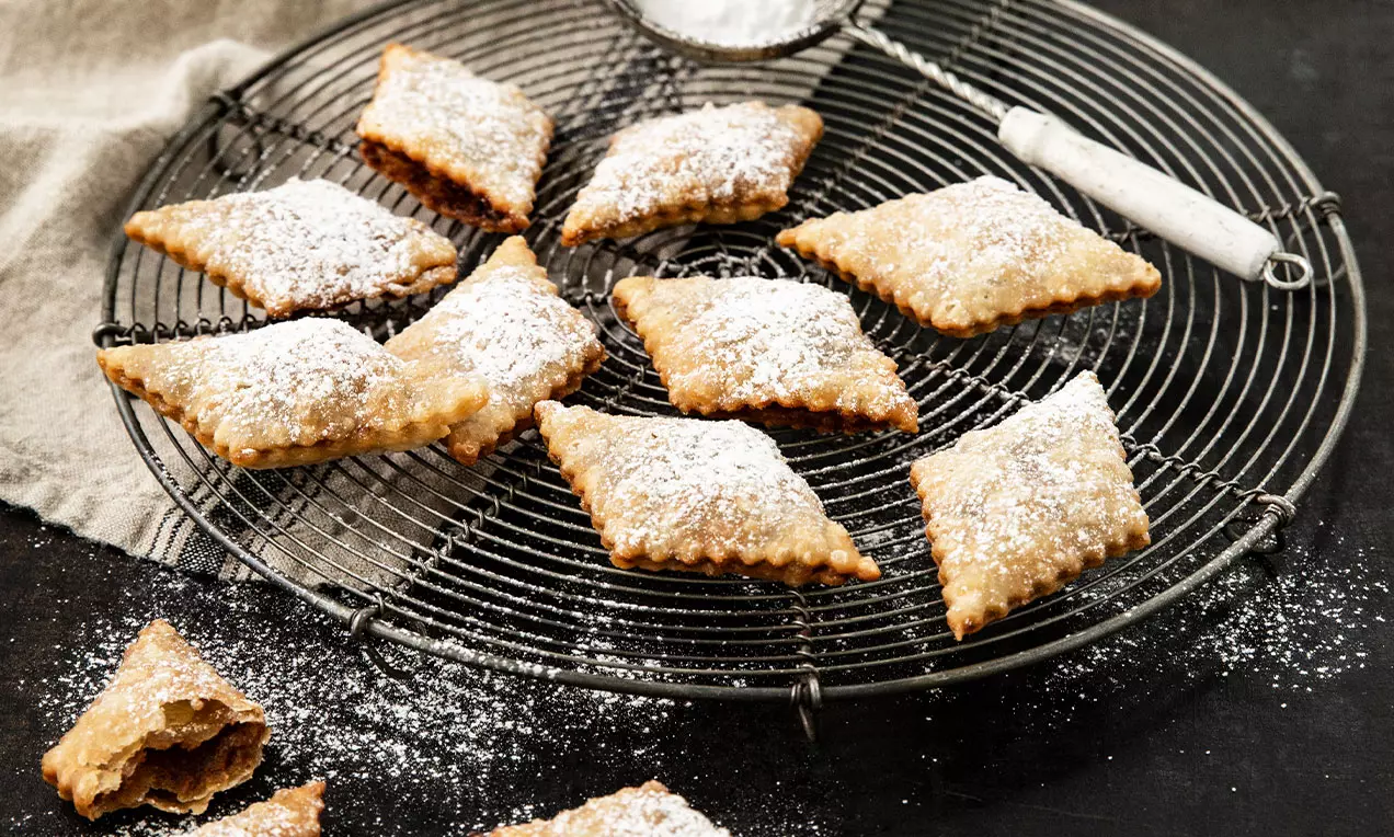 Carrés dorés au bord dentelé légèrement poudrés de sucre glace, disposés sur une grille noire avec une passoire à sucre