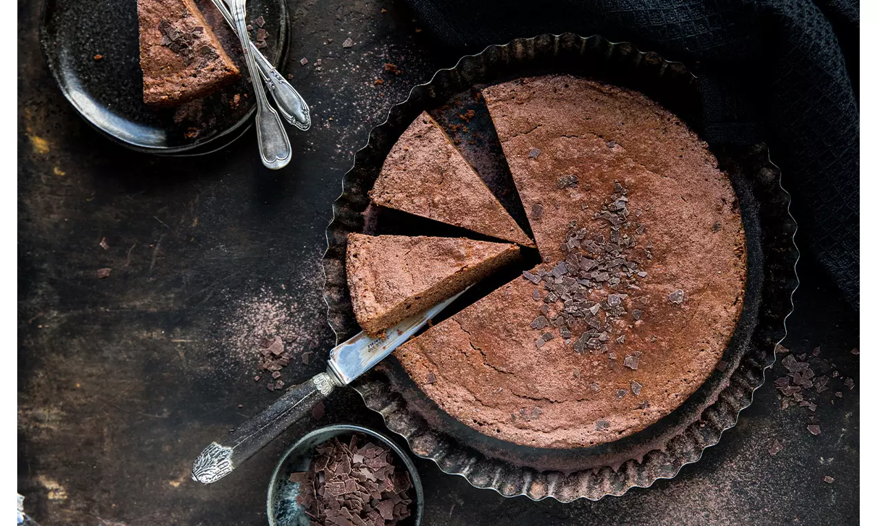 Gâteau rond chocolaté à la texture dense et moelleuse, parsemé d'éclats de chocolat, dans un moule noir avec cuillères argentées