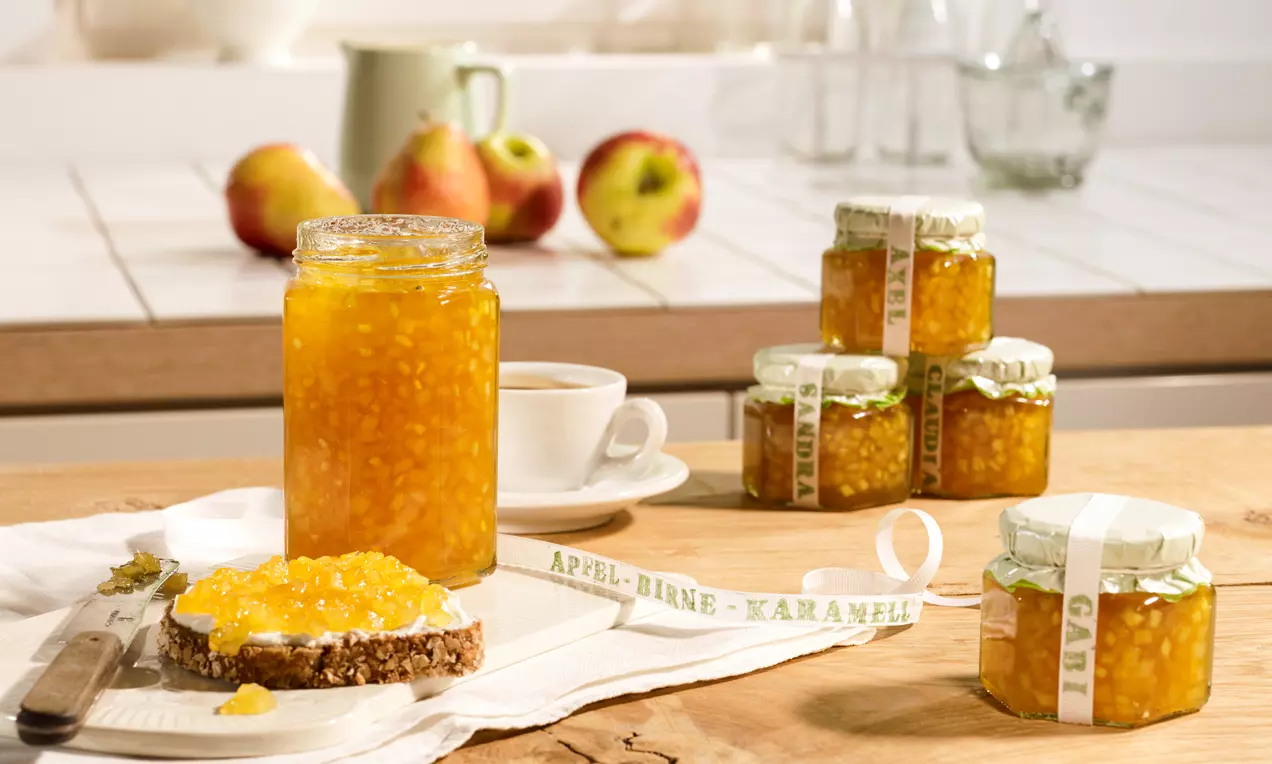 Gläser mit goldgelber Apfel-Birnen-Konfitüre auf Holz, ein offenes Glas neben einer Scheibe Brot mit Konfitüre und Äpfel im Hintergrund