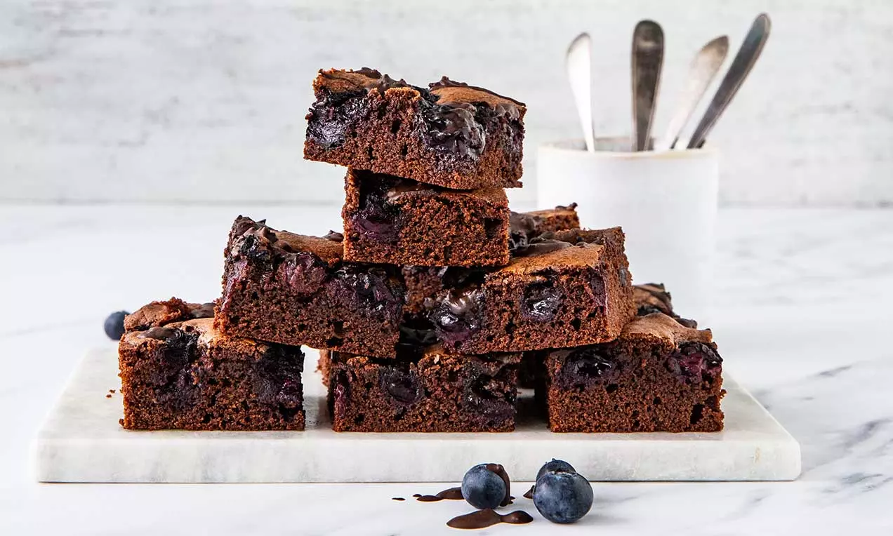 Brownies quadrati dal colore marrone intenso, punteggiati di mirtilli succosi, disposti a piramide su un piatto di marmo bianco con mirtilli freschi accanto
