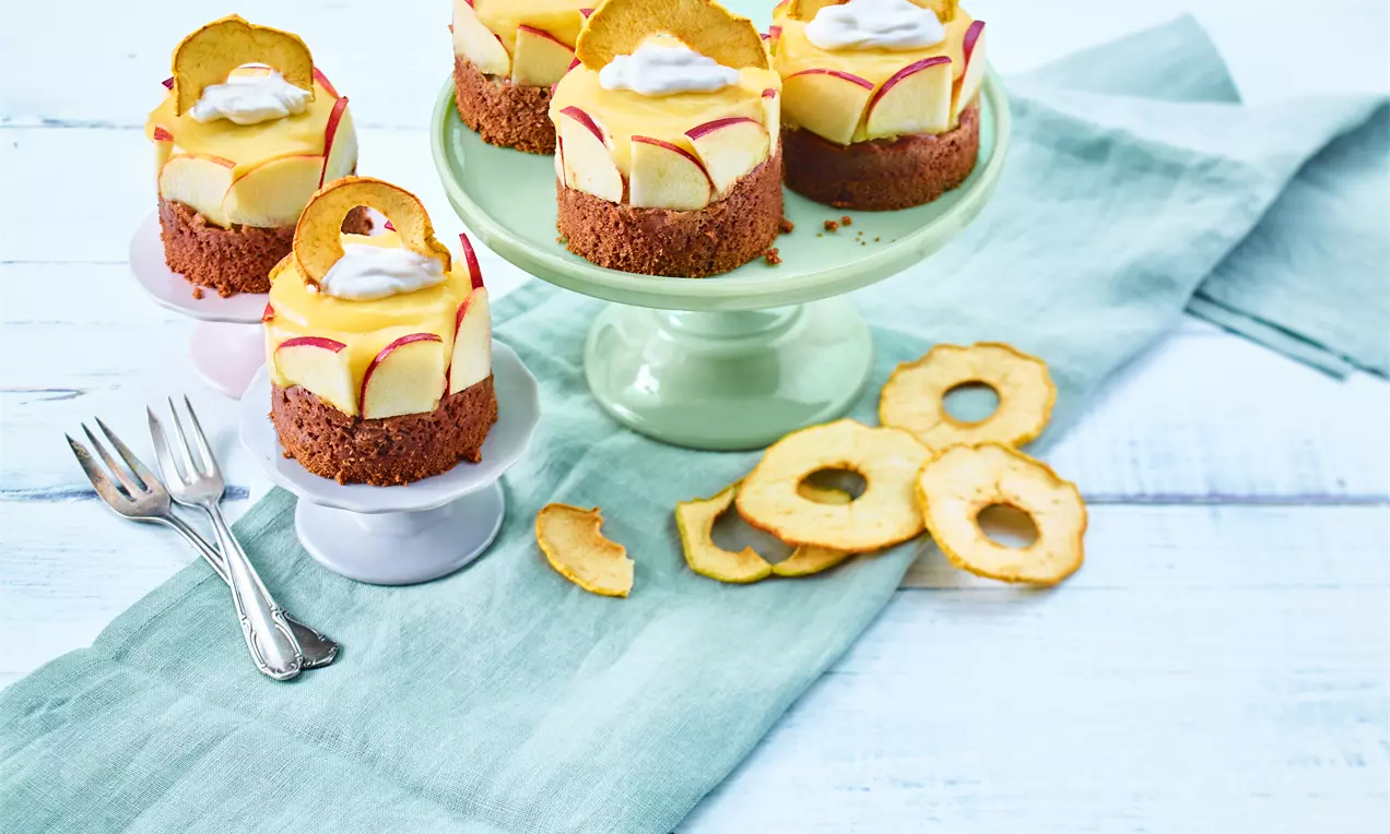 Vegane Apfeltörtchen mit gelber Creme und braunem Boden, dekoriert mit dünnen Apfelscheiben und Sahnetupfen auf grüner Tortenplatte