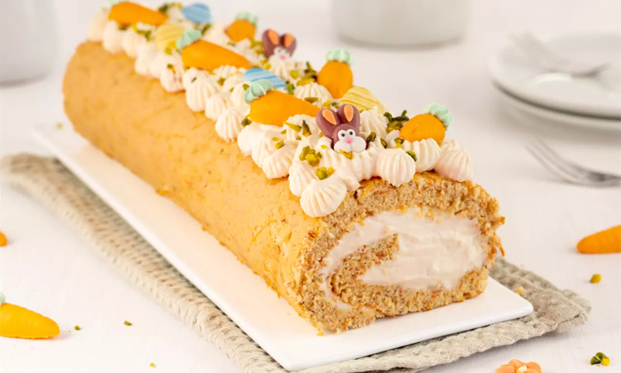 Goldbraune Rüebli-Biskuitrolle mit weißer Quark-Joghurt Füllung, dekoriert mit Sahnetupfen, Marzipanmöhren und Blüten auf weißer Platte