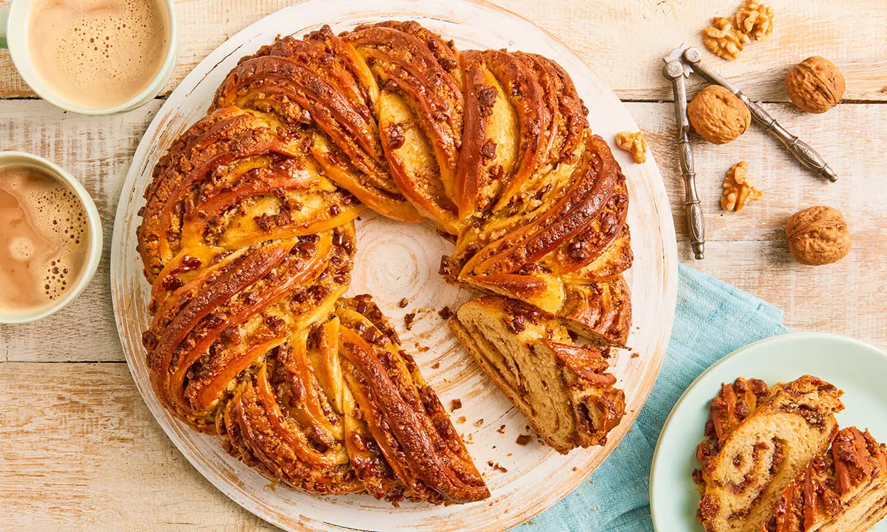 Couronne en pâte levée dorée et brillamment glacée, garnie de noix hachées, présentée sur une assiette avec café et noix adjacents