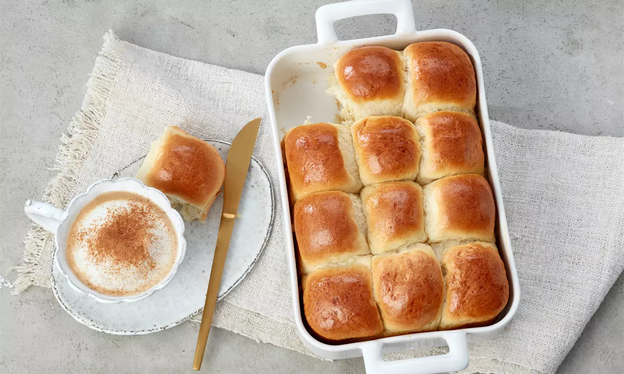 Goldbraune, weich glänzende Milchbrötchen in einer weißen Auflaufform, daneben ein Teller mit Brötchenstück, goldfarbenem Messer und Cappuccino in weißer Tasse