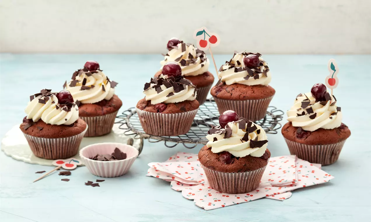 Schwarzwälder Kirsch-Cupcakes mit dunklem Teig, weißer Sahnehaube, Schokoraspeln und Kirschen auf einem Gitter mit Papierdeko angerichtet
