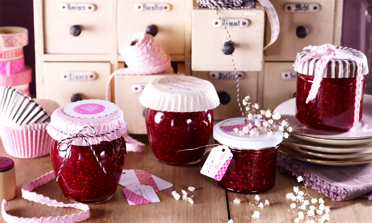 Barattoli di marmellata rosso rubino con coperchi decorati e etichette rosa su tavolo di legno chiaro accanto a piatti e nastri