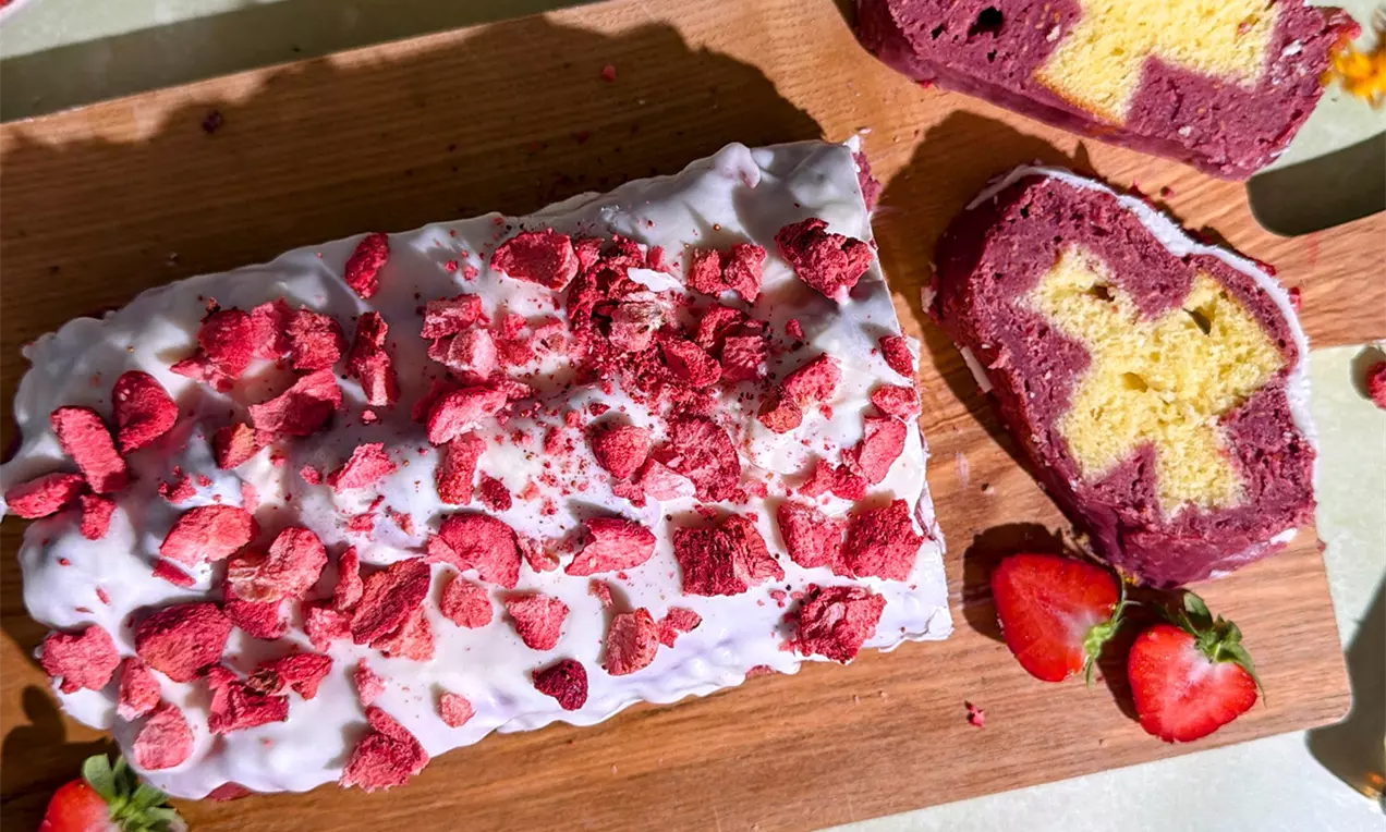 Rechteckiger Kuchen mit weißem Zuckerguss und roten Beerenstücken, daneben herzförmige Stücke mit gelbem Kreuz auf lila Grund, dekoriert mit Erdbeeren