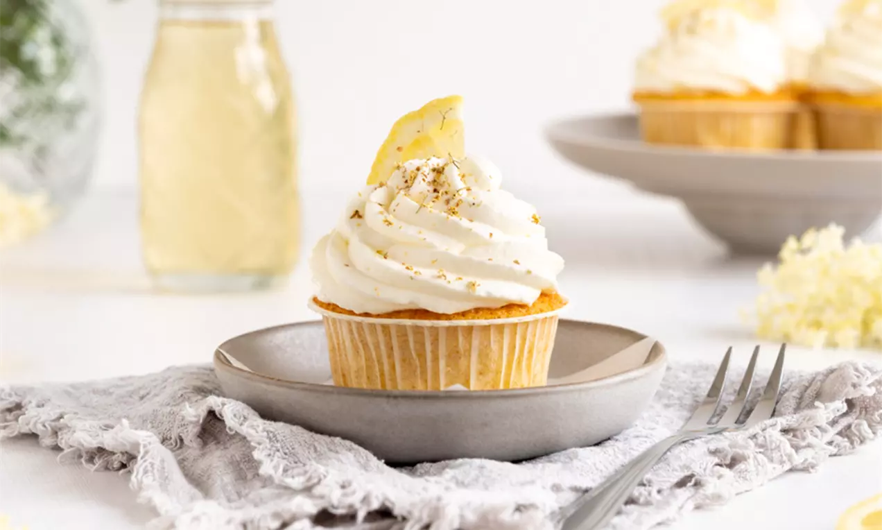Goldgelber Zitronen-Cupcake mit luftiger Holunderblütencreme, garniert mit Zitronenscheibe und Kräutern, auf grauem Teller mit Stoffserviette