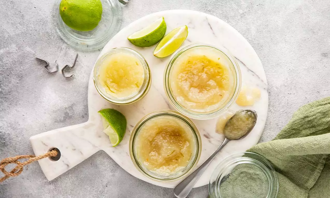 Confettura dorata e lucida in piccoli vasetti di vetro su un tagliere di marmo bianco, con lime tagliati e un cucchiaio argentato accanto