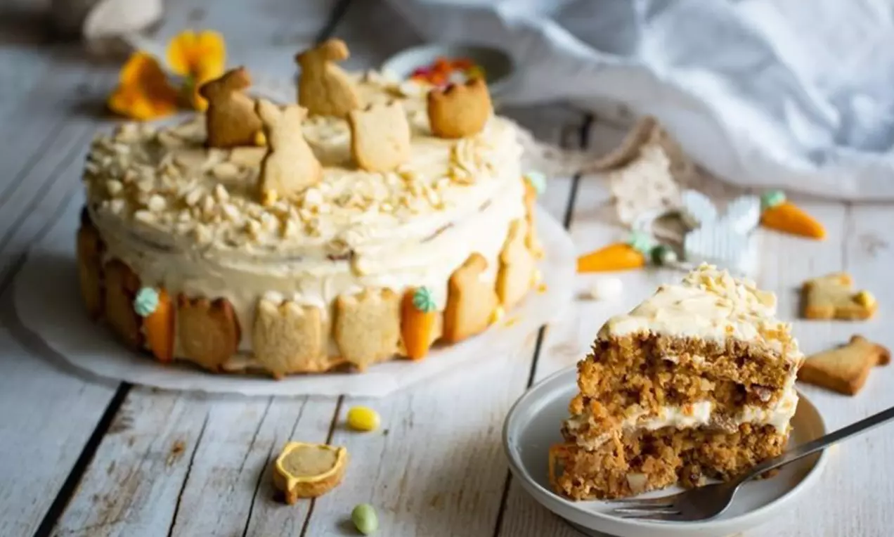 Karottenkuchen mit heller Creme, dekoriert mit Kekshasen und Nüssen, auf Teller angerichtet, umgeben von kleinen Süßigkeiten und Deko-Elementen