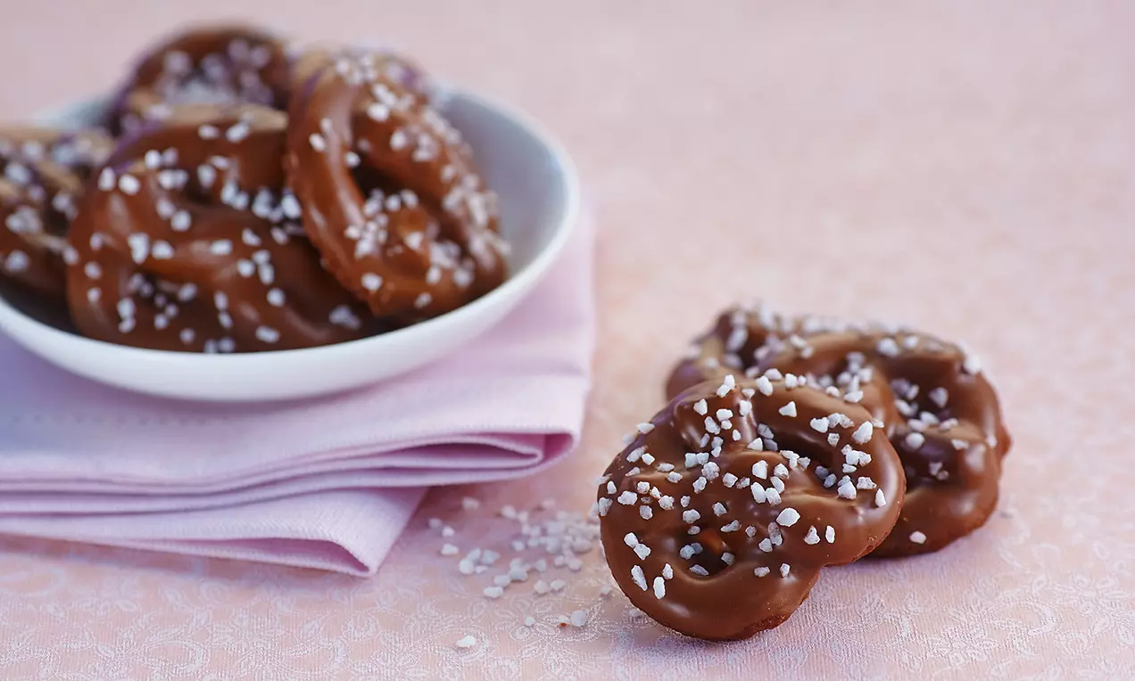 Brezel al cioccolato color marrone lucido ricoperte di granelli di zucchero bianco, disposte su un piatto bianco e tovagliolo rosa