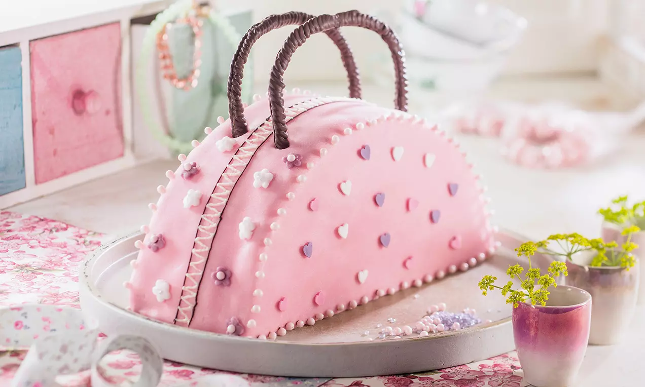 Gâteau en forme de sac à main rose pastel, orné de perles de sucre blanches, de fleurs colorées et de anses au chocolat brun