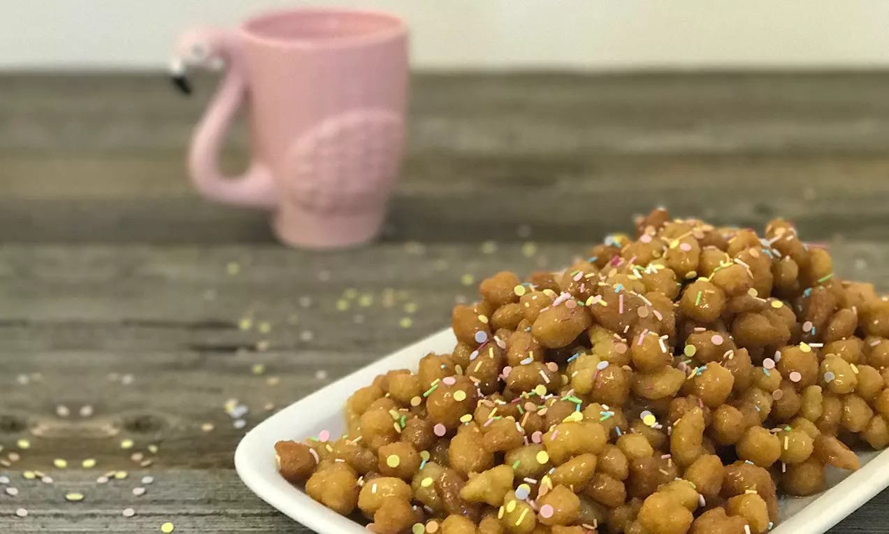 Goldbraune kleine Teigkügelchen mit glänzendem Überzug und bunten Streuseln auf weißer Platte, dahinter ein rosa Becher auf Holztisch
