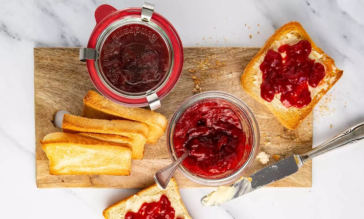 Confiture rouge rubis à la fraise et à la vanille dans des pots transparents, étalée sur des tranches de pain dorées sur une planche en bois