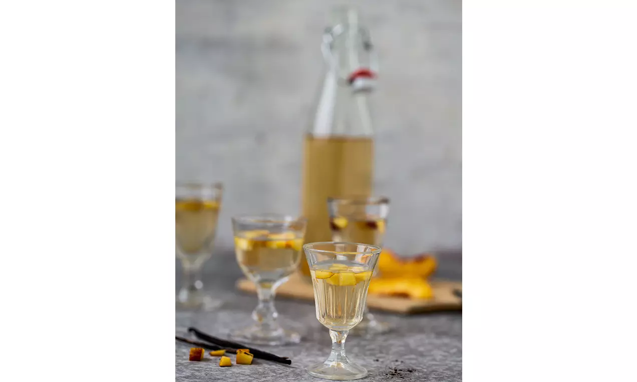 Klarer Aprikosen-Pfirsich-Likör in kleinen Gläsern mit gelben Fruchtwürfeln, dazu Flasche und frische Fruchtstücke auf grauem Untergrund