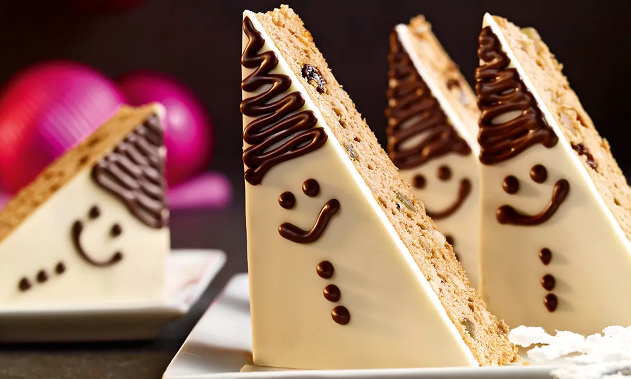 Fette triangolari ricoperte di glassa bianca, decorate con dettagli di cioccolato a forma di volti sorridenti e capelli ricurvi su fondo scuro