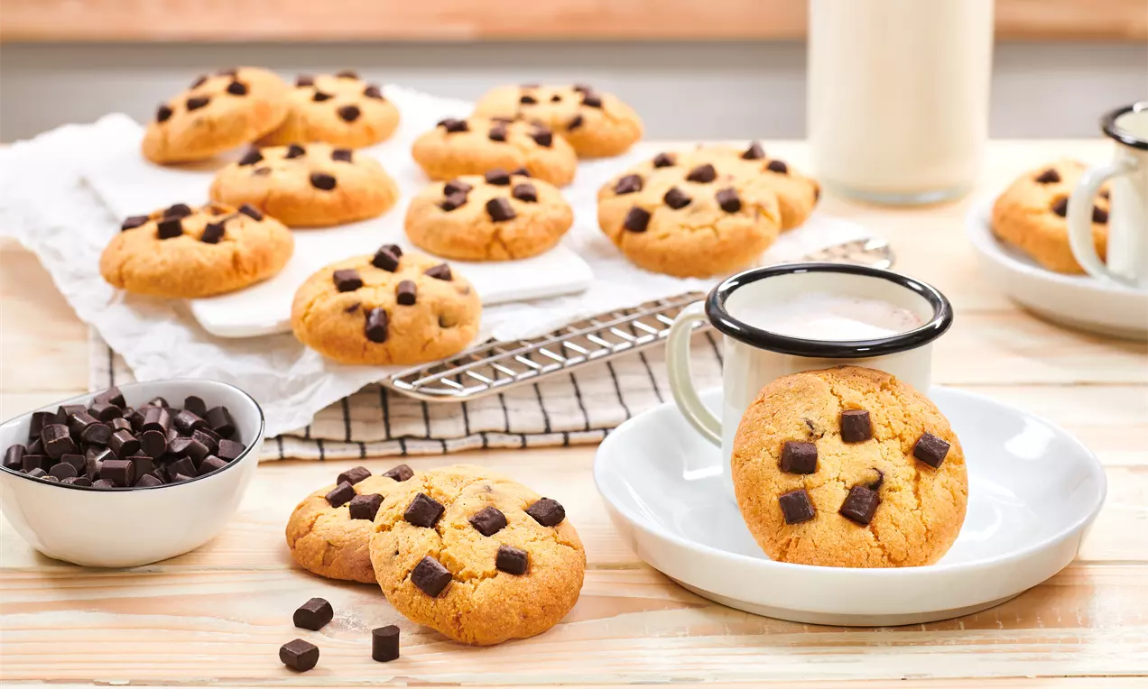 Cookies dorés à la surface légèrement craquelée, parsemés de cubes de chocolat noir, sur grille, assiette blanche et bois clair